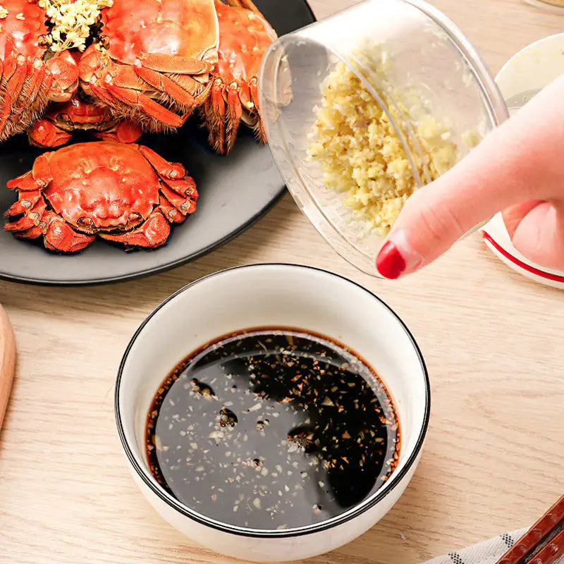 Crabs on a plate with a bowl of dark sauce and a hand holding a small container of garlic on a wooden table.