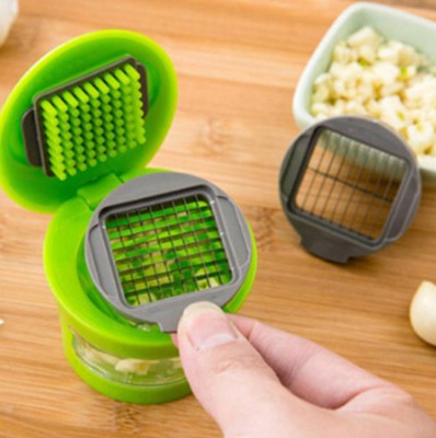 Green and gray garlic press on a wooden surface with garlic and a bowl of minced garlic in the background.