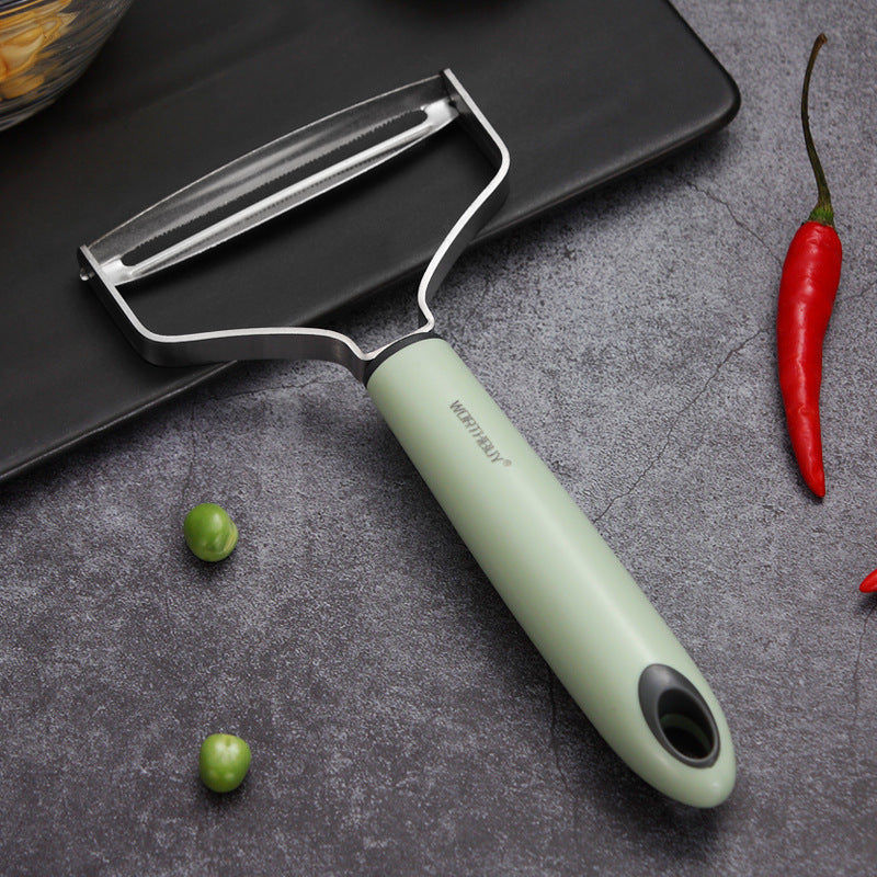 Y-shaped vegetable peeler with a green handle on a gray surface with vegetables.