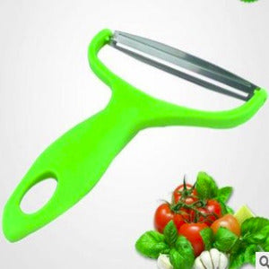 A bright green handheld vegetable peeler with a stainless steel blade, shown on a white background with tomatoes, basil, and garlic at the bottom right.
