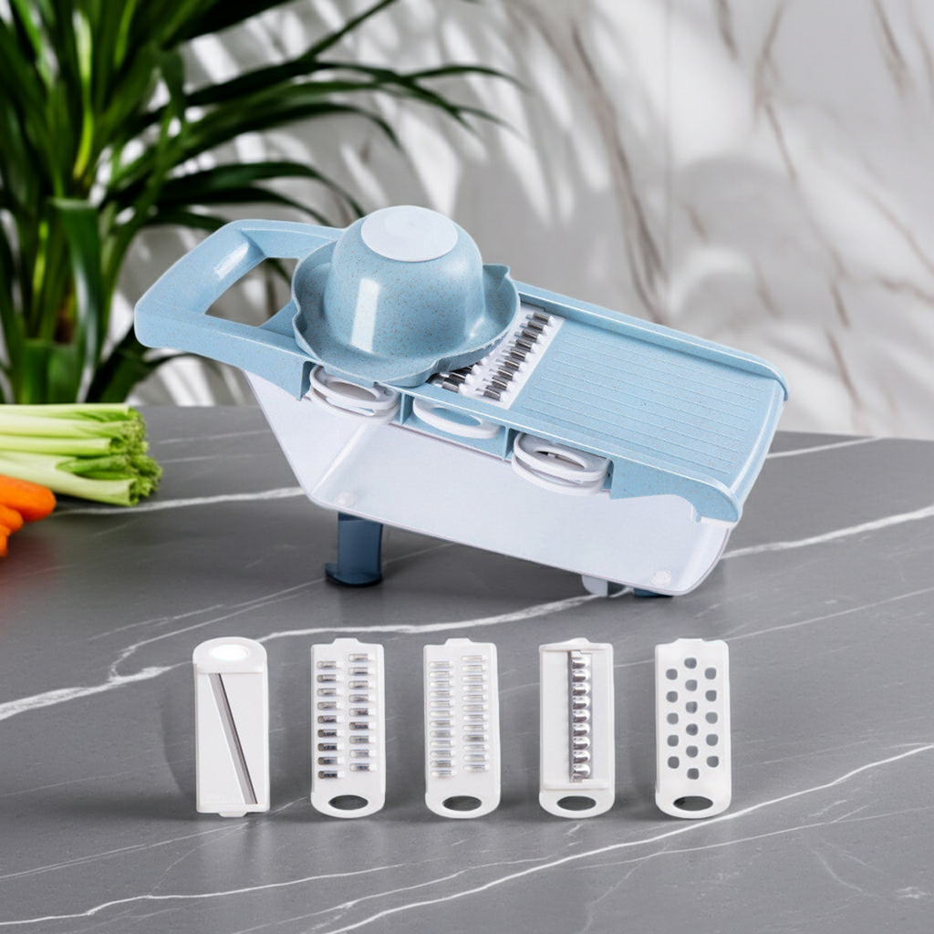 A kitchen mandoline/slicer attachment set displayed on a gray marble countertop, with the main light blue and white slicer body, a bowl insert, and four interchangeable blade attachments arranged in front. Vegetables and a plant are visible in the background as styling props.