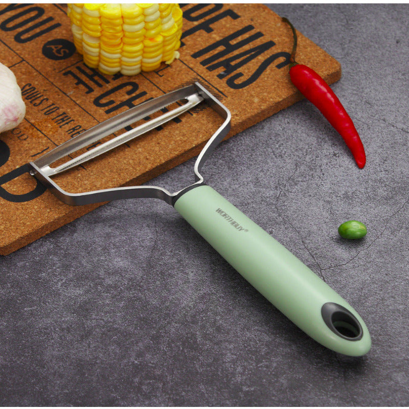 Green vegetable peeler on a gray surface with kitchen items like corn, red chili, and green bean.