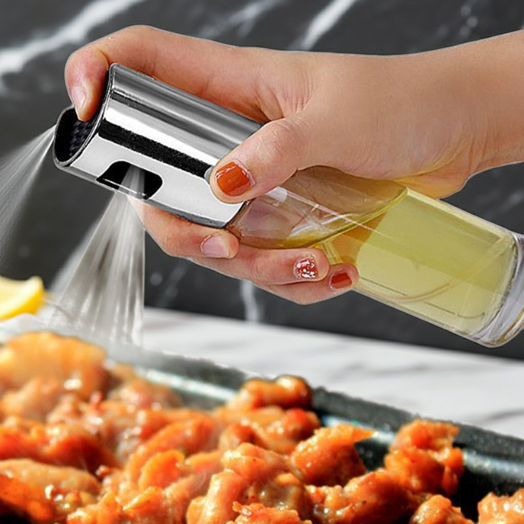 A hand holding a clear glass spray bottle with a shiny silver metal nozzle, spraying oil over cooked food on a dark tray. The background is a dark marble-patterned surface.