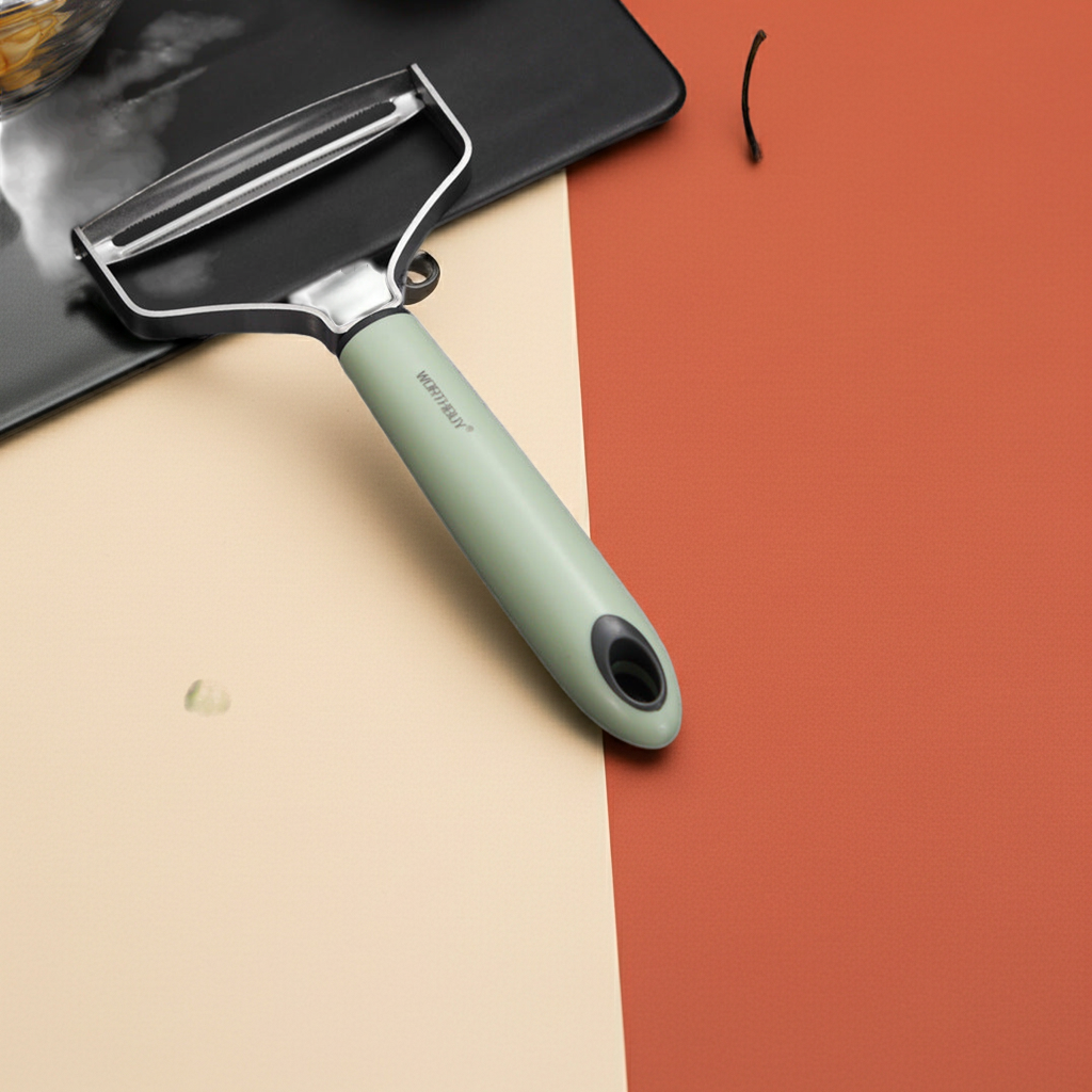 A kitchen peeler with a sage green handle and silver metal blade, shown diagonally on a split beige and terracotta background.