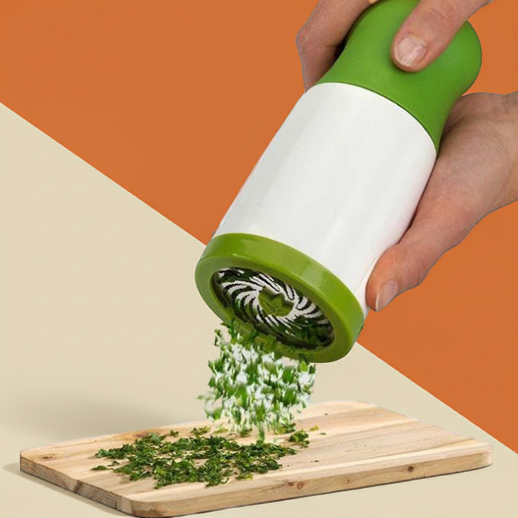 A hand holds a green and white manual herb chopper over a wooden cutting board, with chopped green herbs falling from the spout onto the board against an orange and beige background.
