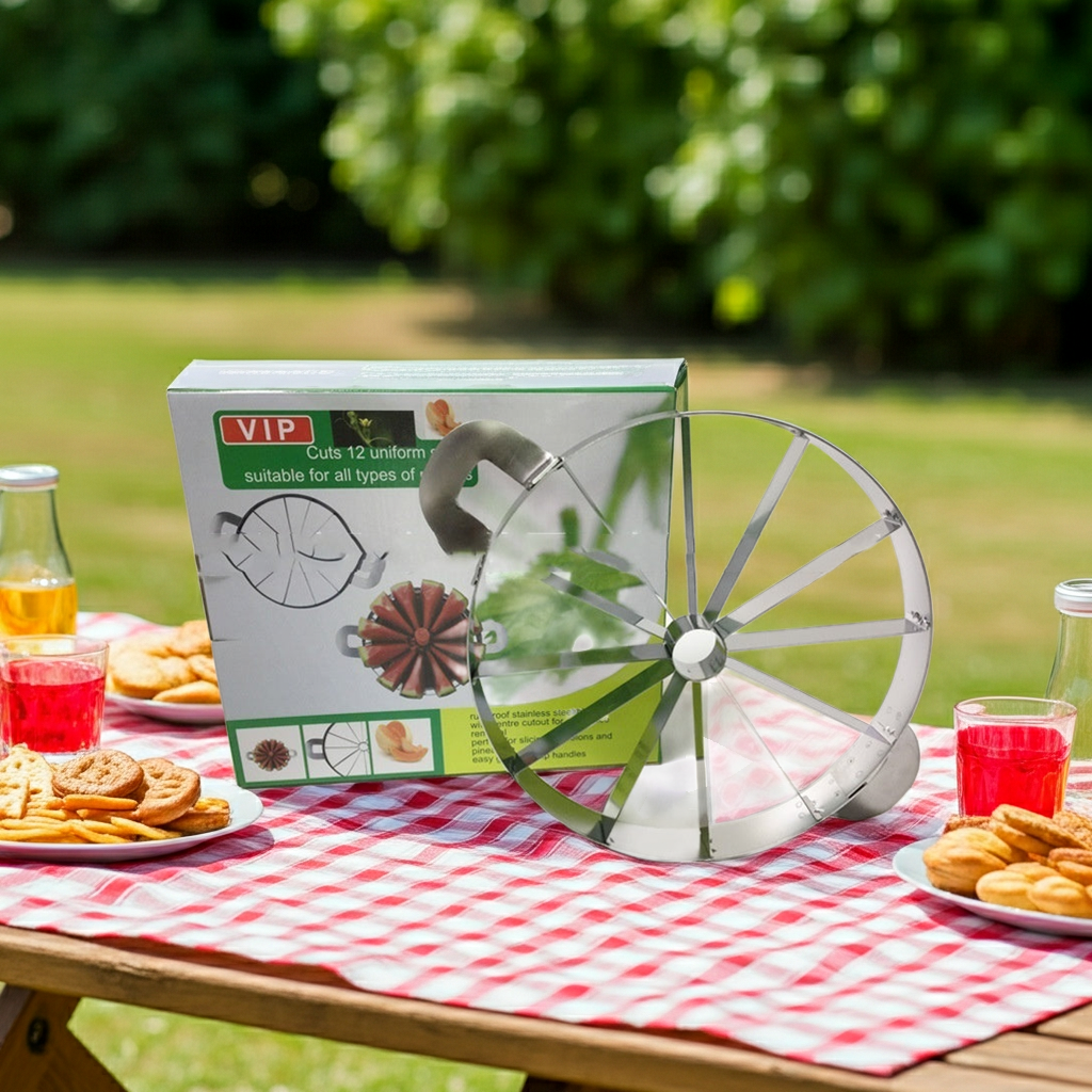 A metal 12-slice pizza cutter wheel displayed next to its retail packaging on a red-and-white checkered picnic table outdoors, with cookies, drinks, and a blurred green garden background.