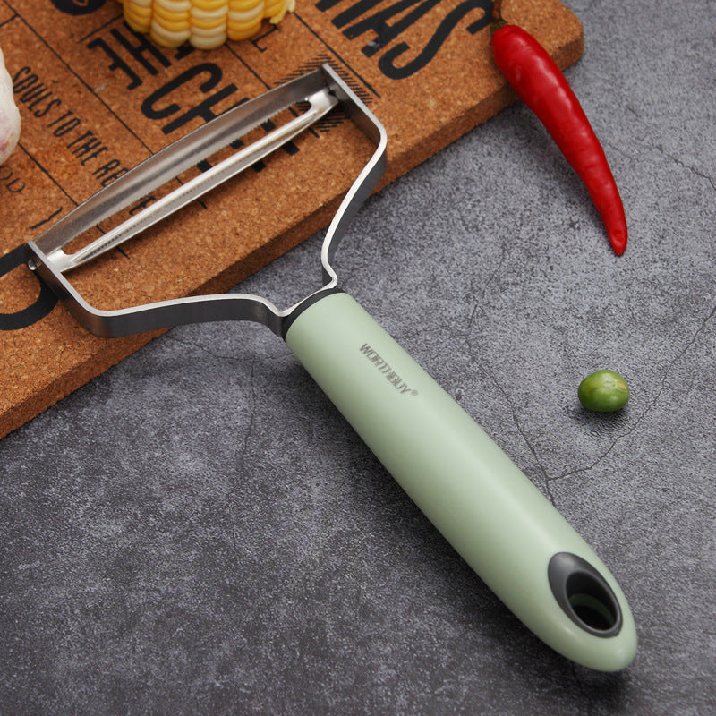 Green vegetable peeler on a gray surface with a cutting board and vegetables in the background