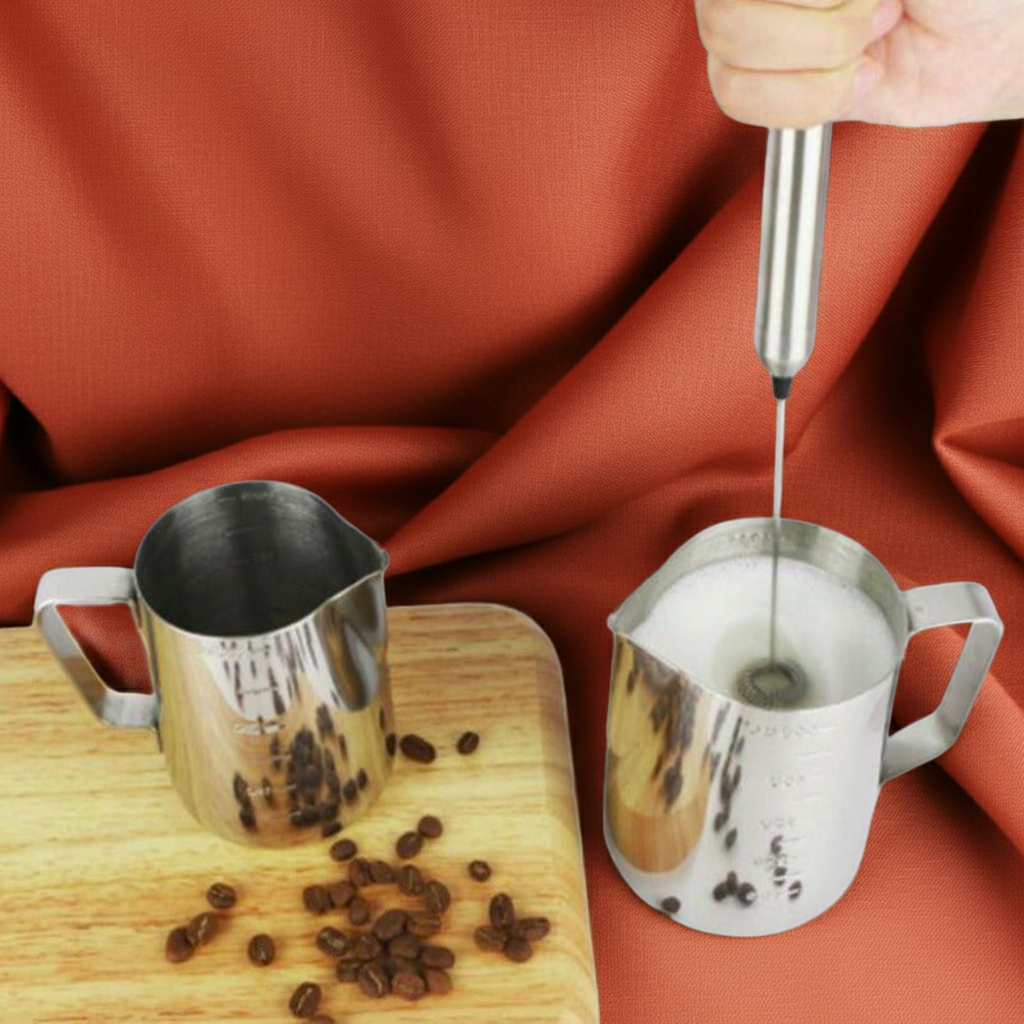 A stainless steel milk frother is being used to foam milk in a metal pitcher, with another metal pitcher and scattered coffee beans on a wooden board against a draped rust-orange fabric background.