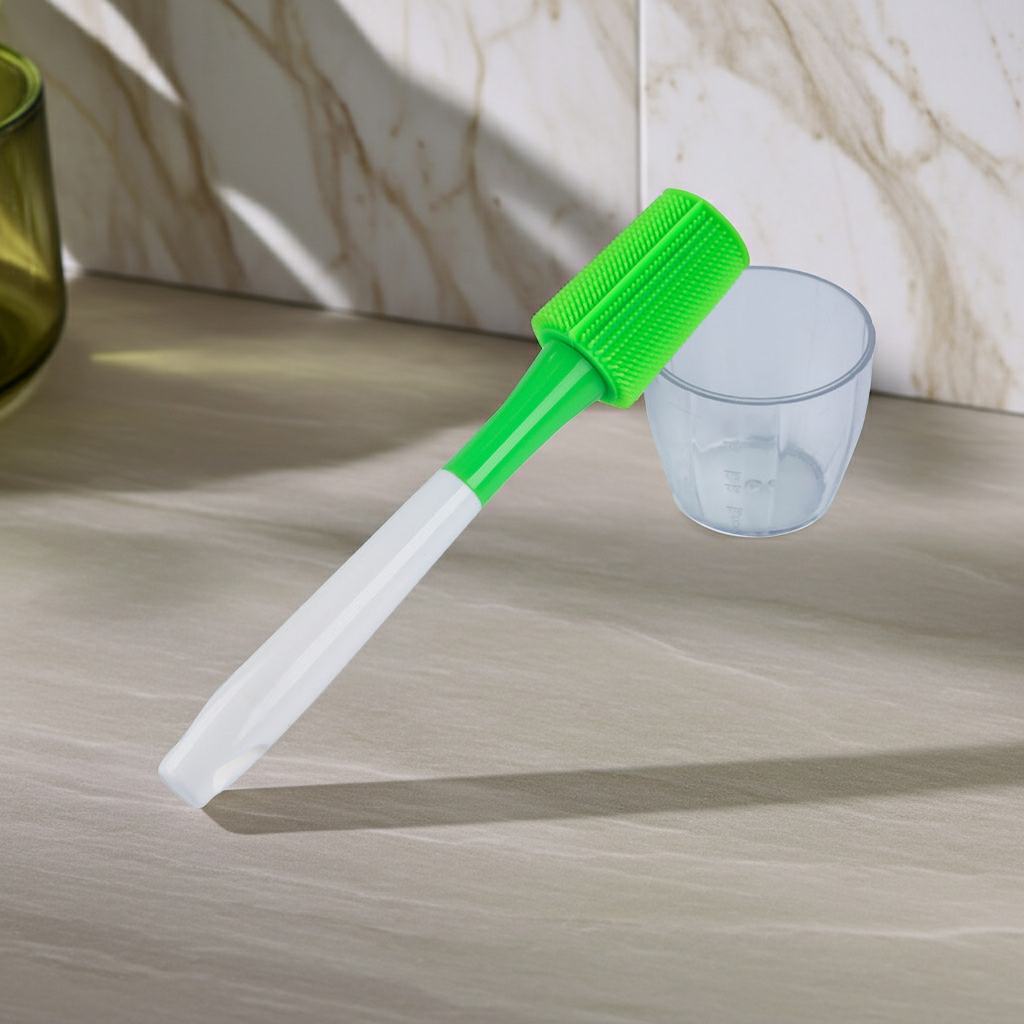 A silicone cleaning brush with a bright green bristle head and a translucent white handle, shown next to a small clear measuring cup on a beige marble countertop.