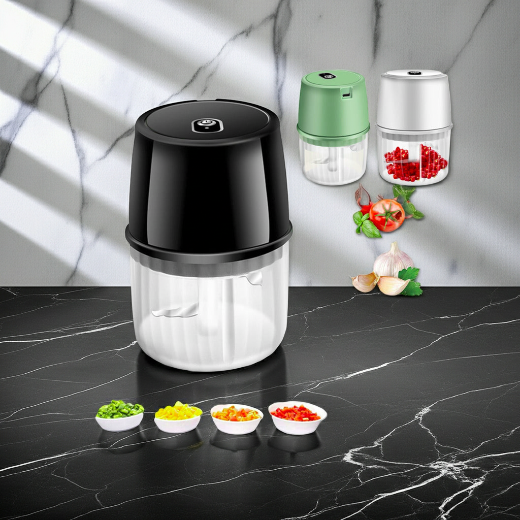 A countertop electric food chopper shown in black and clear, with smaller green and silver versions in the background. The main unit sits on a dark marble surface, with chopped vegetables and garlic arranged nearby against a marble wall backdrop.