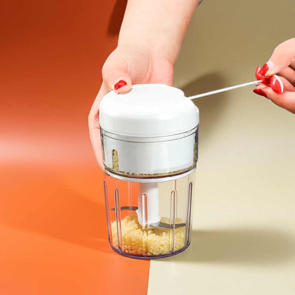 A small manual food chopper with a white top lid and clear container, being held by a hand with red nail polish while another hand pushes a pin through the top. The chopper contains minced garlic, set against a split orange and beige background.