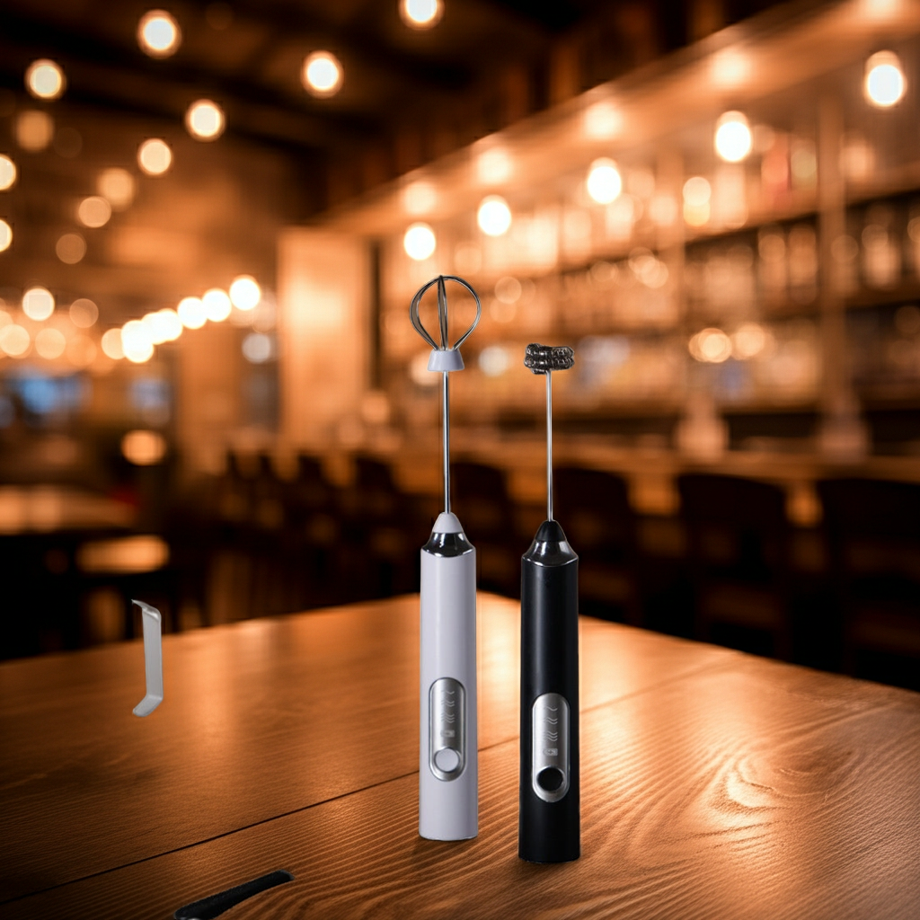 Two handheld electric milk frothers standing on a wooden table in a warmly lit bar or cafe setting. One frother is white with a silver whisk attachment, and the other is black with a matching attachment.
