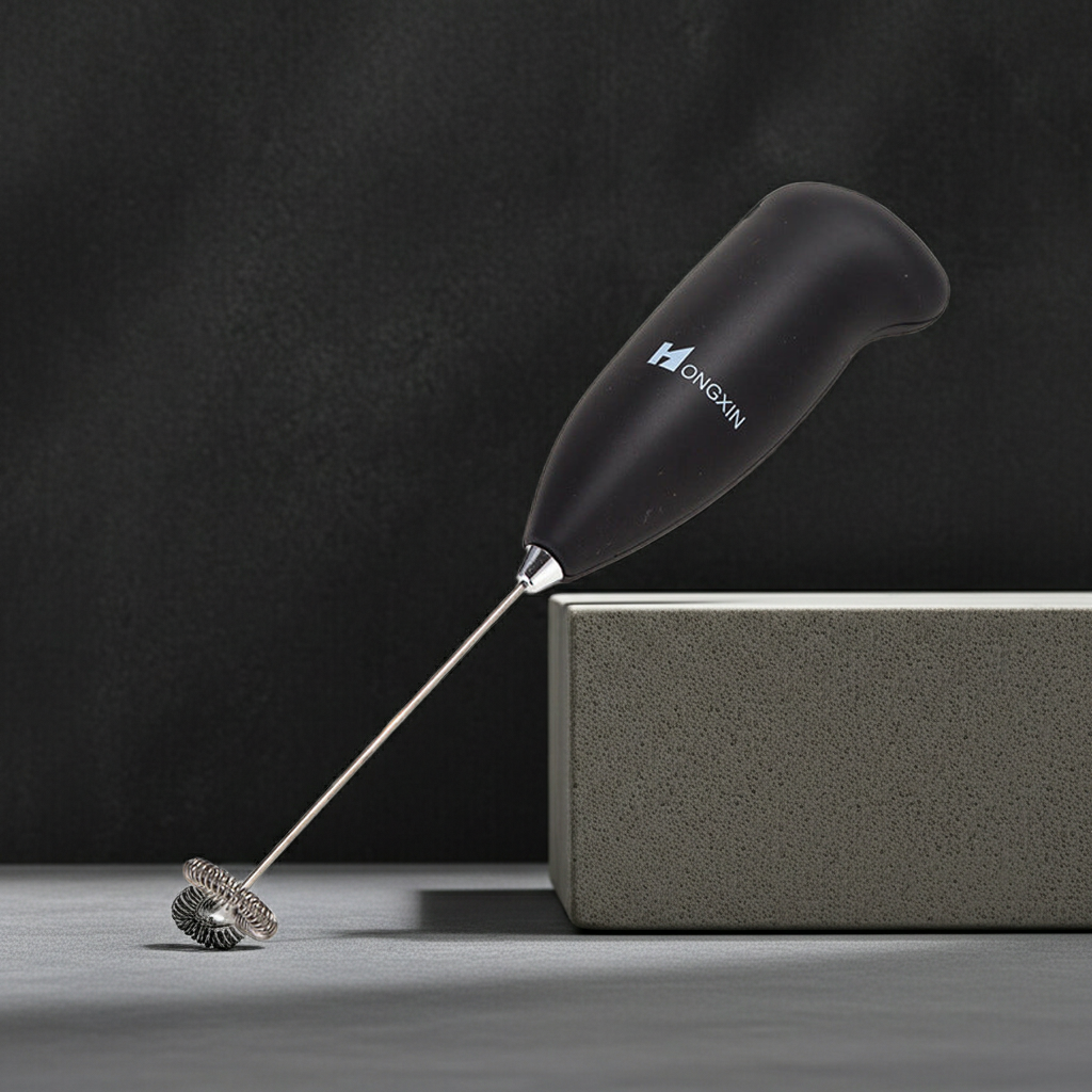 A handheld milk frother with a matte black handle and a silver metal whisker, resting on a gray block against a dark textured background. The handle features the brand name 'GANGYUAN' in white.